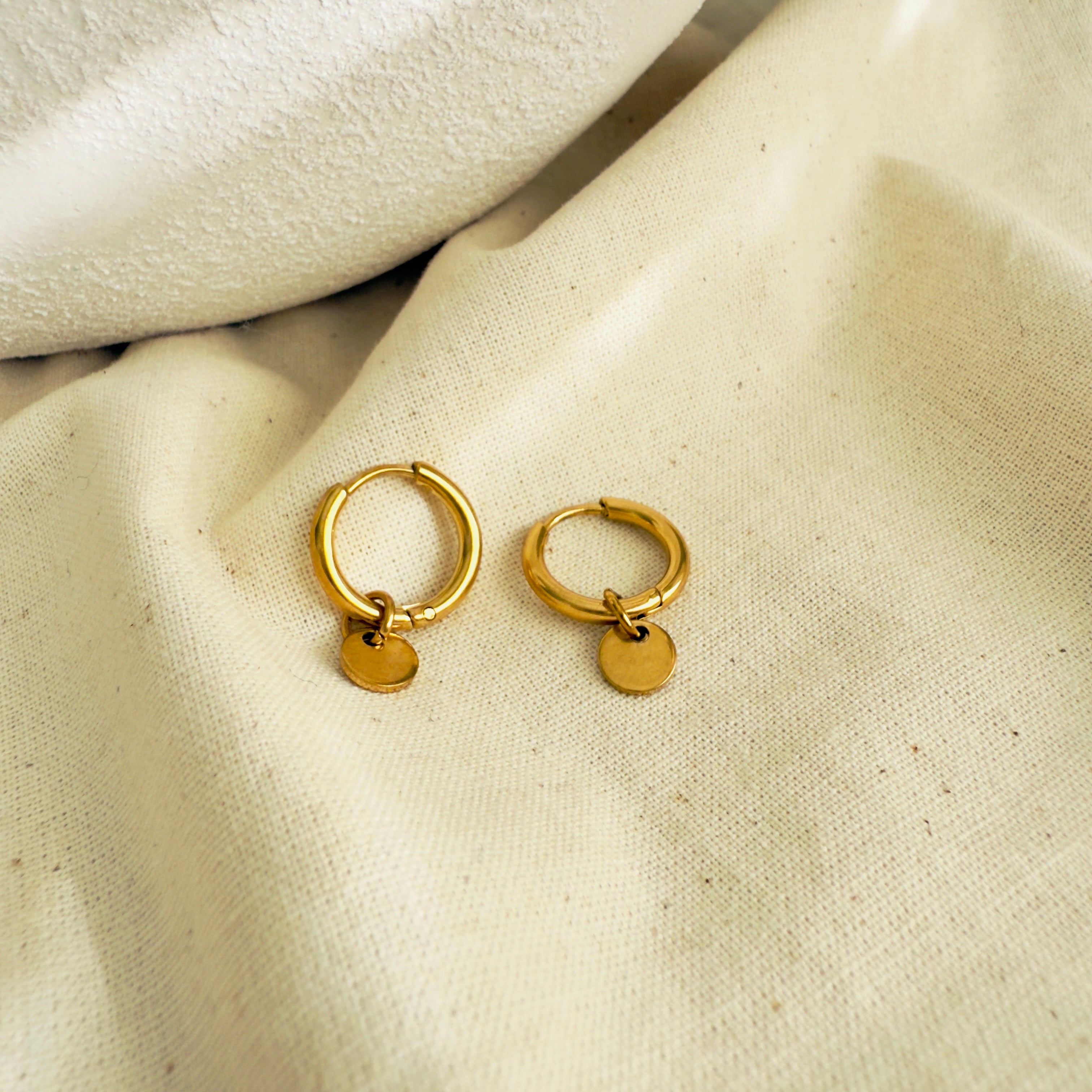 Gold hoop earrings with small round tags on a beige fabric background