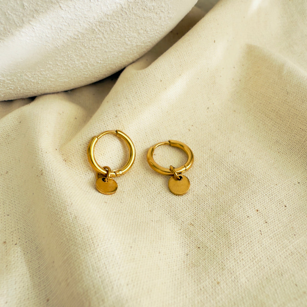 Gold hoop earrings with small round tags on a beige fabric background